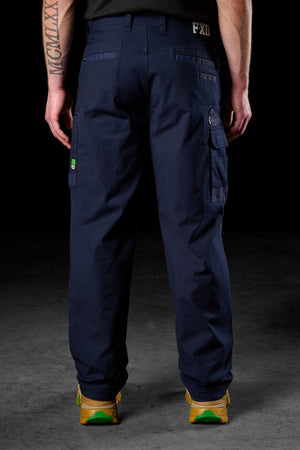 A person faces away from the camera in FXD WP-3L Loose Fit Stretch Canvas Work Pants (navy blue), paired with yellow-green shoes. Tattooed arms are visible against a dark background.