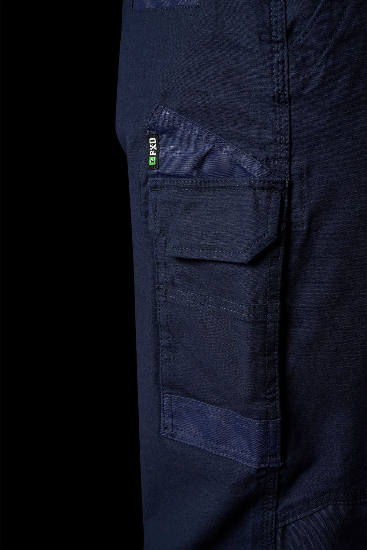 Close-up of FXD WP-3L Loose Fit Stretch Canvas Work Pants in dark blue, featuring side pockets with flaps, visible stitching, and a small black and green PXG tag near the upper pocket.