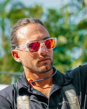 A man with long hair pulled back is wearing SafeStyle Fusions Clear Frame Mirror Red Polarised sunglasses and a black shirt with orange earbuds around his neck. He is outdoors, surrounded by trees and a clear blue sky.