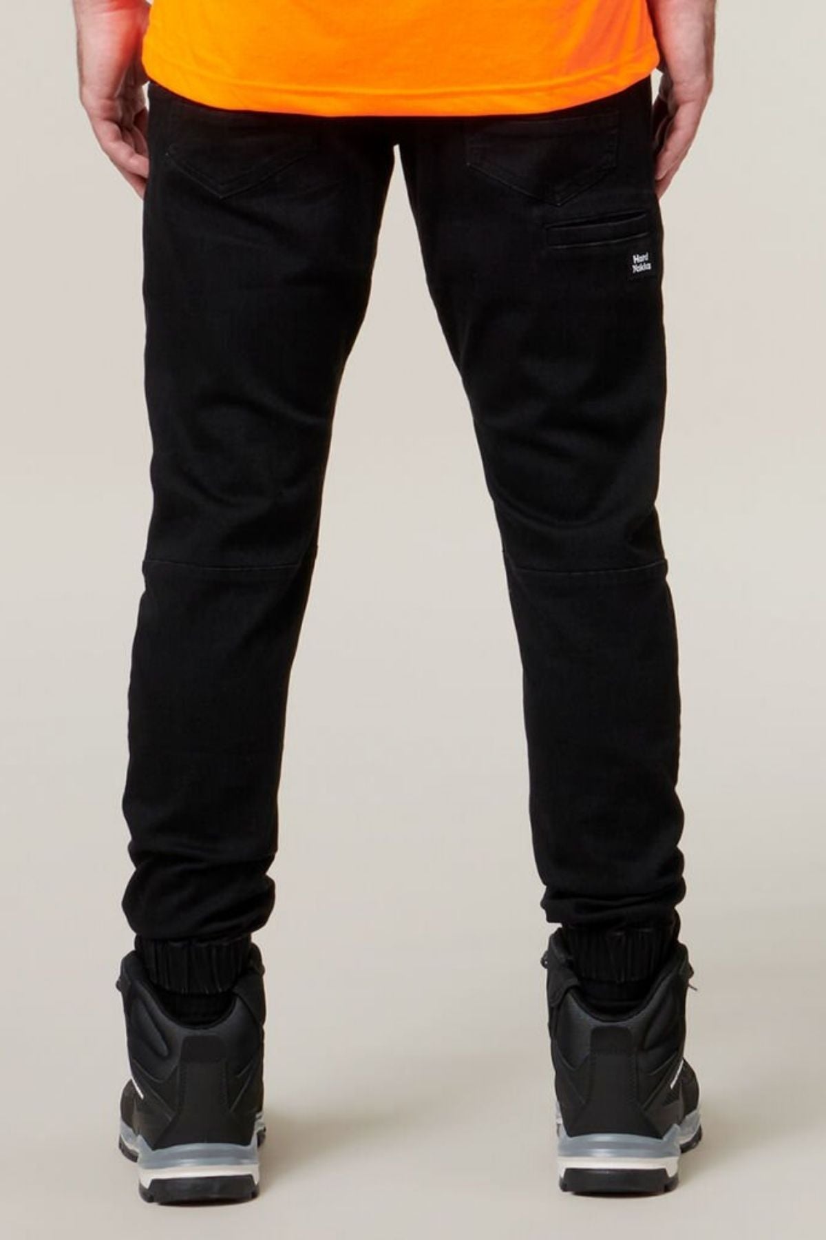 A person wearing Hard Yakka Active Denim Jogger Pants with a cargo pocket, black sneakers with white soles, and an orange shirt is photographed from the back. The background is plain and light-colored.