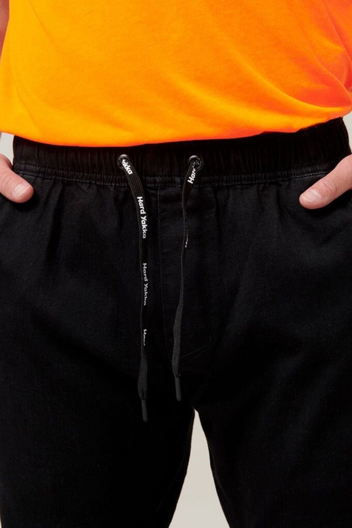Close-up of a person wearing Hard Yakka Active Denim Jogger Pants with their hands in their pockets. The pants, featuring a cargo pocket and elastic waist, have a black drawstring with white text. Only the bottom portion of the person's orange shirt is visible.