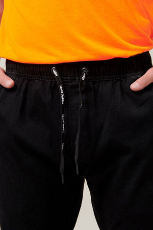 Close-up of a person wearing Hard Yakka Active Denim Jogger Pants with their hands in their pockets. The pants, featuring a cargo pocket and elastic waist, have a black drawstring with white text. Only the bottom portion of the person's orange shirt is visible.