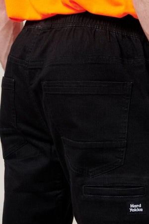 A person wearing Hard Yakka Active Denim Jogger Pants in black, featuring visible back pockets and an elastic waist, is shown. They are also wearing an orange top, and the "Hard Yakka" logo is visible at the bottom right corner of the pants.