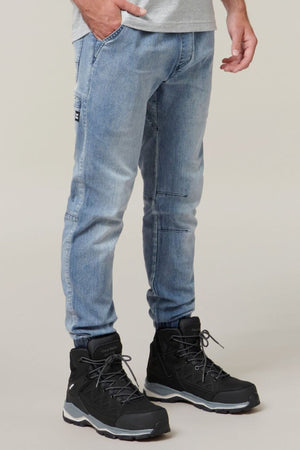A person is shown from the waist down, wearing Hard Yakka Active Denim Jogger Pants in light blue and black hiking boots with gray accents and laces. One hand is in their pocket, exuding a sense of comfortable wear, and the background is plain and neutral.
