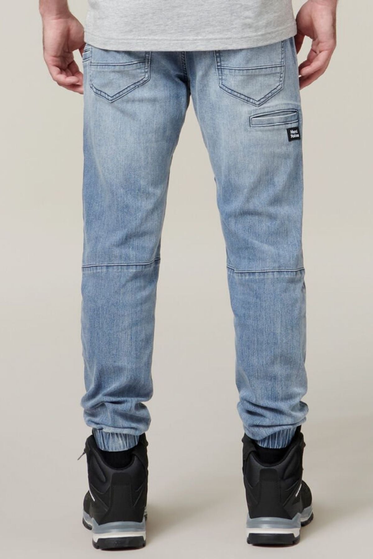 Rear view of a person wearing light blue fitted Hard Yakka Active Denim Jogger Pants, a grey shirt, and black sneakers with white soles. The pants feature two back pockets, an additional vertical cargo pocket on the right side, and elasticated cuffs at the ankles. The background is neutral and plain.