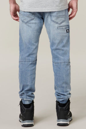 Rear view of a person wearing light blue fitted Hard Yakka Active Denim Jogger Pants, a grey shirt, and black sneakers with white soles. The pants feature two back pockets, an additional vertical cargo pocket on the right side, and elasticated cuffs at the ankles. The background is neutral and plain.