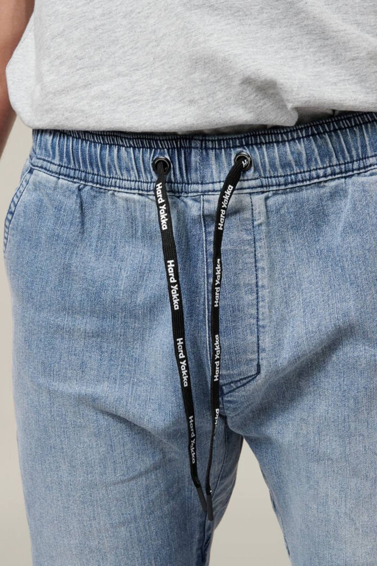 Close-up of a person wearing the Hard Yakka Active Denim Jogger Pant in light blue, featuring an elastic waistband and black drawstrings with white "Hard Yakka" text. The individual, dressed for comfort, also sports a light gray shirt. Their hands are partially visible, resting casually near the pockets.