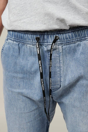 Close-up of a person wearing the Hard Yakka Active Denim Jogger Pant in light blue, featuring an elastic waistband and black drawstrings with white "Hard Yakka" text. The individual, dressed for comfort, also sports a light gray shirt. Their hands are partially visible, resting casually near the pockets.