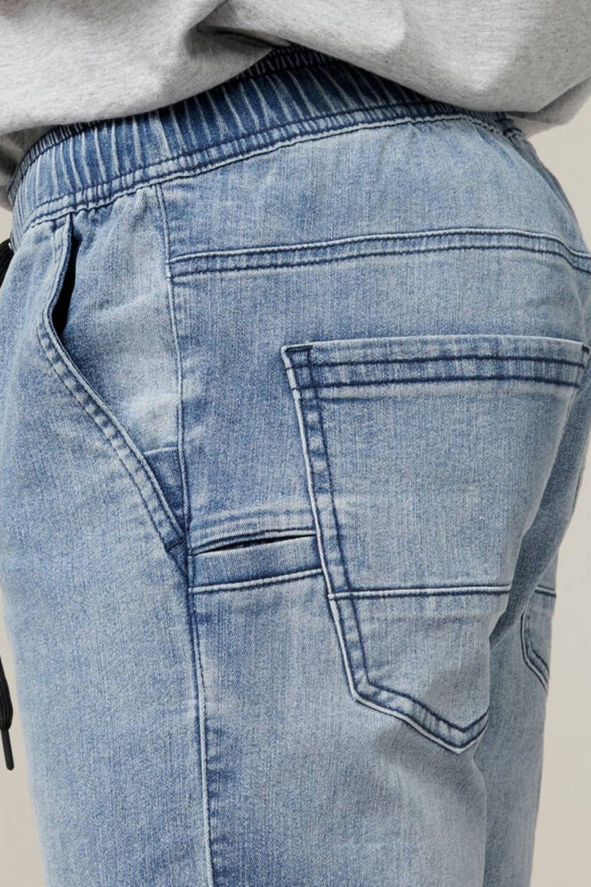 Close-up side view of a person wearing the Hard Yakka Active Denim Jogger Pant in light blue, which features an elastic waistband. The comfortable design includes a large back pocket with visible stitching and appears slightly loose-fitting. The person is also wearing a light gray top.