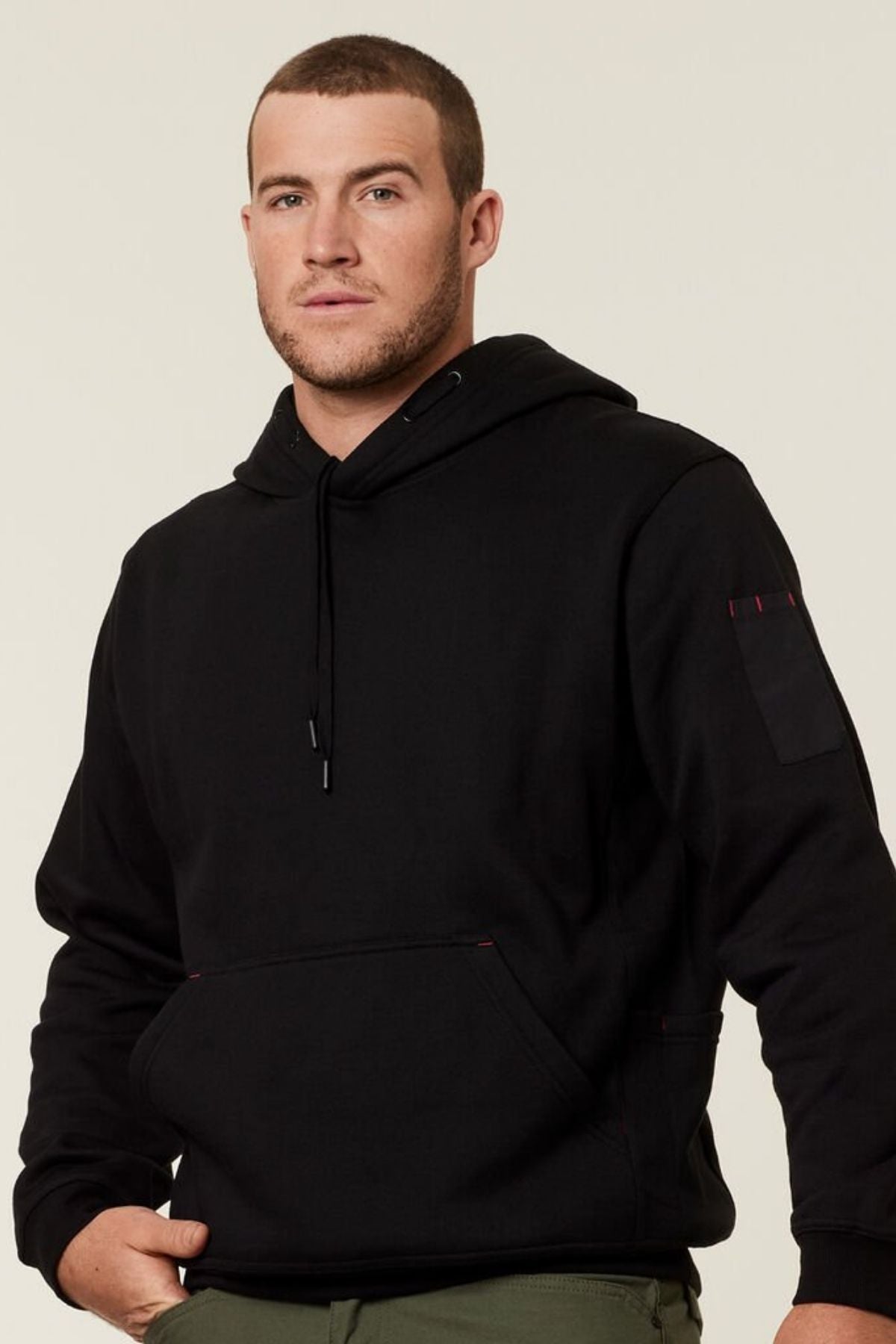 A man with short hair, dressed in a Hard Yakka Plain Fleece Hoodie, stands against a plain background. With his hands tucked into the front pocket of the brushed fleece hoodie, he looks straight at the camera with a neutral expression.