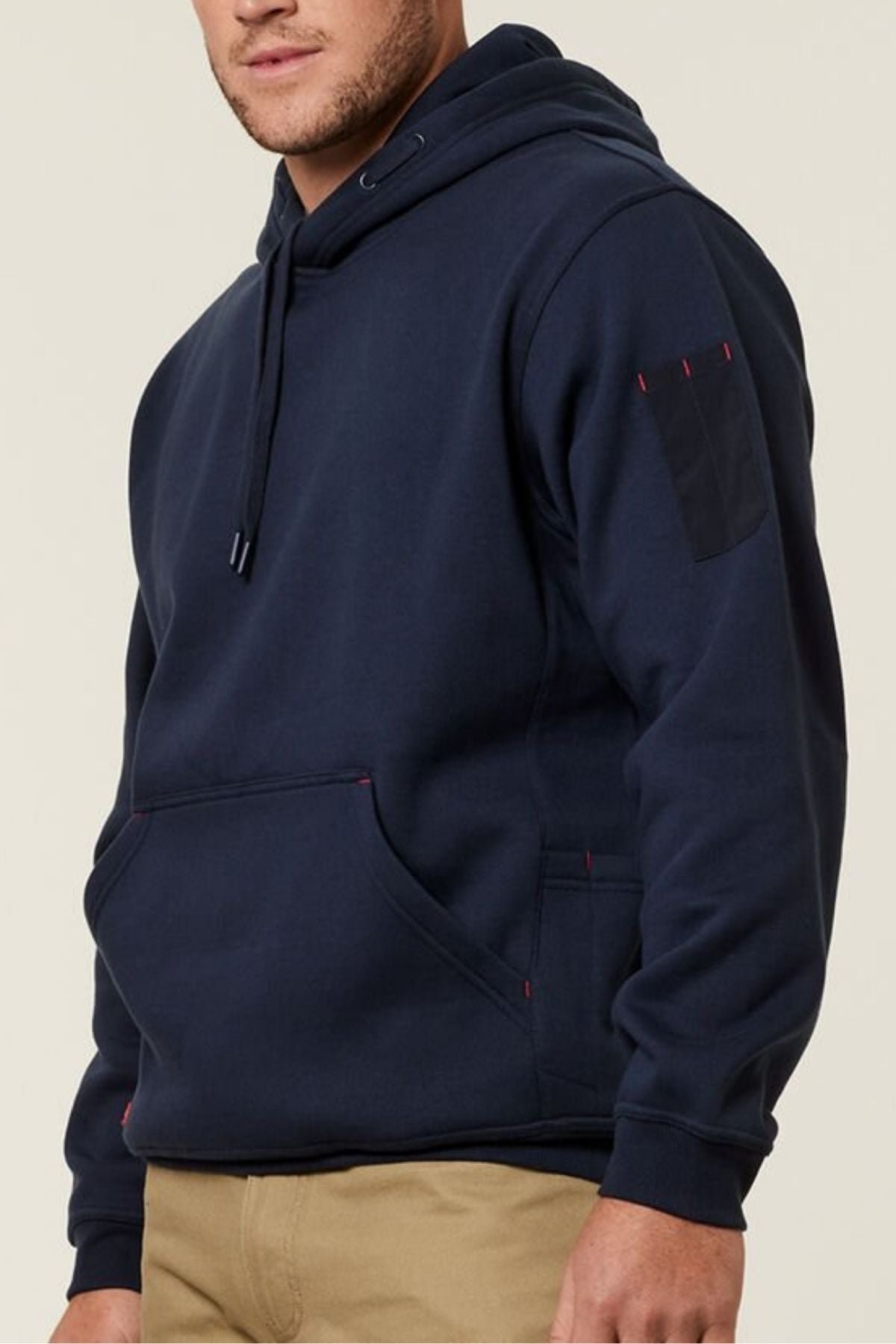Against a plain background, someone stands wearing a Hard Yakka Plain Fleece Hoodie in dark navy featuring reinforced stitching, a kangaroo pocket, and an upper sleeve pocket. It's paired with light brown pants for stylish worksite functionality.