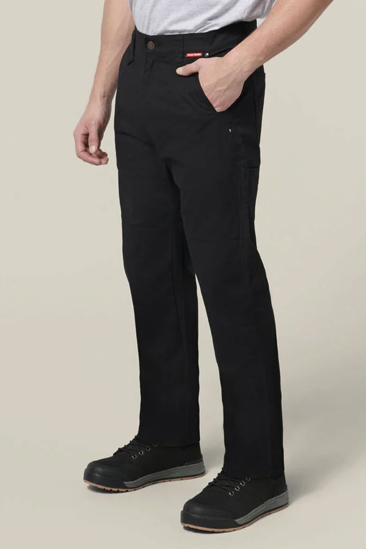 A person models black Hard Yakka Heritage Carpenters Pant with black work shoes, standing against a plain light gray background—one hand in their pocket, embodying classic workwear style.