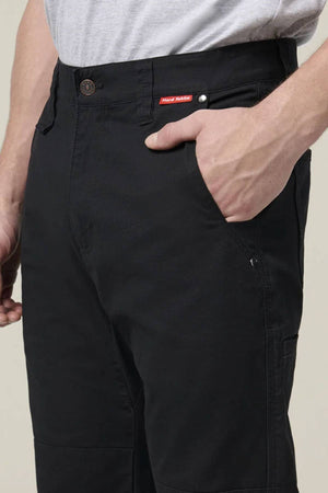 A person wearing Hard Yakka Heritage Carpenters Pant and a light gray shirt stands with their right hand in the front pocket. The black pants have a red and white tag above the pocket.