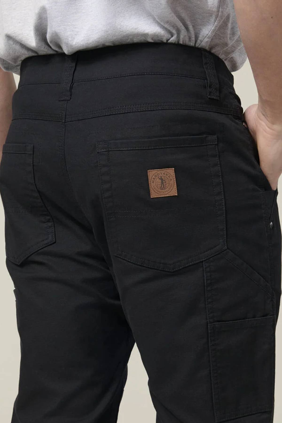 A person wears Hard Yakka Heritage Carpenters Pant, featuring black fabric, visible back pockets, and a brown leather patch—shown waist down with hand on hip—ideal for a rugged workwear look.