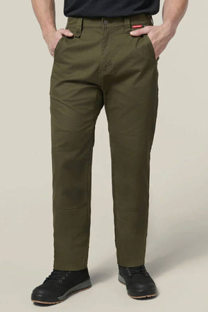 A person wearing olive green Hard Yakka Heritage Carpenters Pant, a black shirt, and black boots stands with hands in pockets against a plain light background. A red tag is visible on the pants near the right pocket.