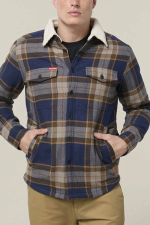 A man stands with hands in pockets, wearing a blue, brown, and gray plaid Hard Yakka Heritage Trucker Sherpa Jacket with a white sherpa collar over a black shirt and tan pants—a classic pick for any winter wardrobe.