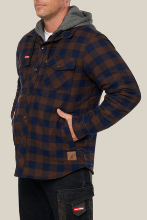 Wearing the Hard Yakka Quilted Flannelette Shacket in brown and navy blue plaid over a gray hooded sweatshirt, a person stands with hands in pockets against a plain beige background.