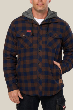 A man in a blue and brown plaid Hard Yakka Quilted Flannelette Shacket with two chest pockets, gray hood with black drawstrings, and black pants stands with one hand in his pocket against a plain background.