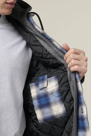 A person opens a Hard Yakka Quilted Flannelette Shacket, revealing a blue and white plaid inner pocket. This cozy winter jacket with 3M Insulation is worn over a light gray shirt.