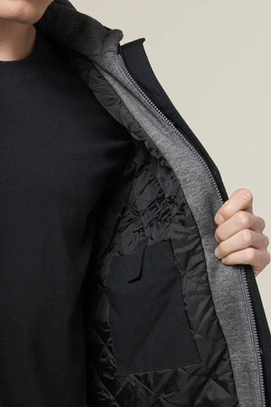 A person in a black shirt opens a grey Hard Yakka Quilted Solid Shacket, showing its quilted black interior and inside pocket. The shacket features durable ripstop outer fabric. The background is plain and light-colored.