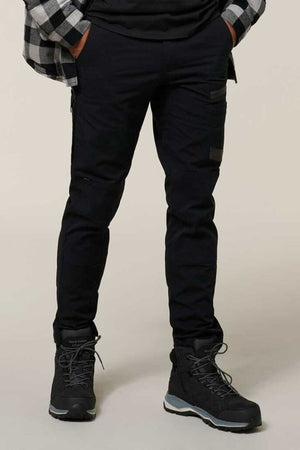A person stands with hands in pockets, wearing black Hard Yakka Raptor Pants, a black shirt, a black and gray plaid shirt, and black hiking boots against a plain light background.
