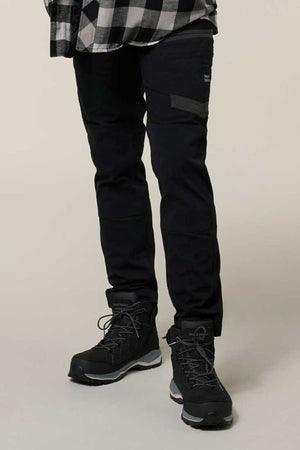 A person in black Hard Yakka Raptor Pant with Repreve ECO Ripstop Technology, black hiking boots, and a black-and-white plaid shirt stands on a light surface; the photo is cropped below the shoulders.