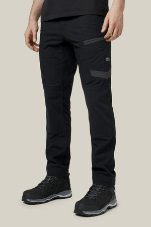 A person wears Hard Yakka Raptor Pant hiking pants with zippered pockets and black hiking shoes, standing against a plain beige background. Only the lower half of their body is visible.