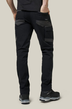 Wearing Hard Yakka Raptor Pant work pants with multiple pockets and black sneakers, a person stands facing away from the camera with one hand in their pocket against a plain light background. Features Repreve ECO Ripstop Technology.