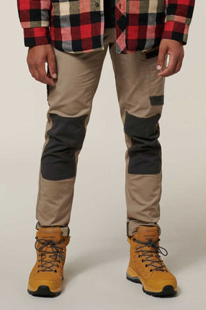 A person is standing wearing a red and black checkered jacket, tan Hard Yakka Raptor Pants with reinforced knee patches featuring Repreve ECO Ripstop Technology, and yellow hiking boots with blue laces. The background is plain and light-colored.