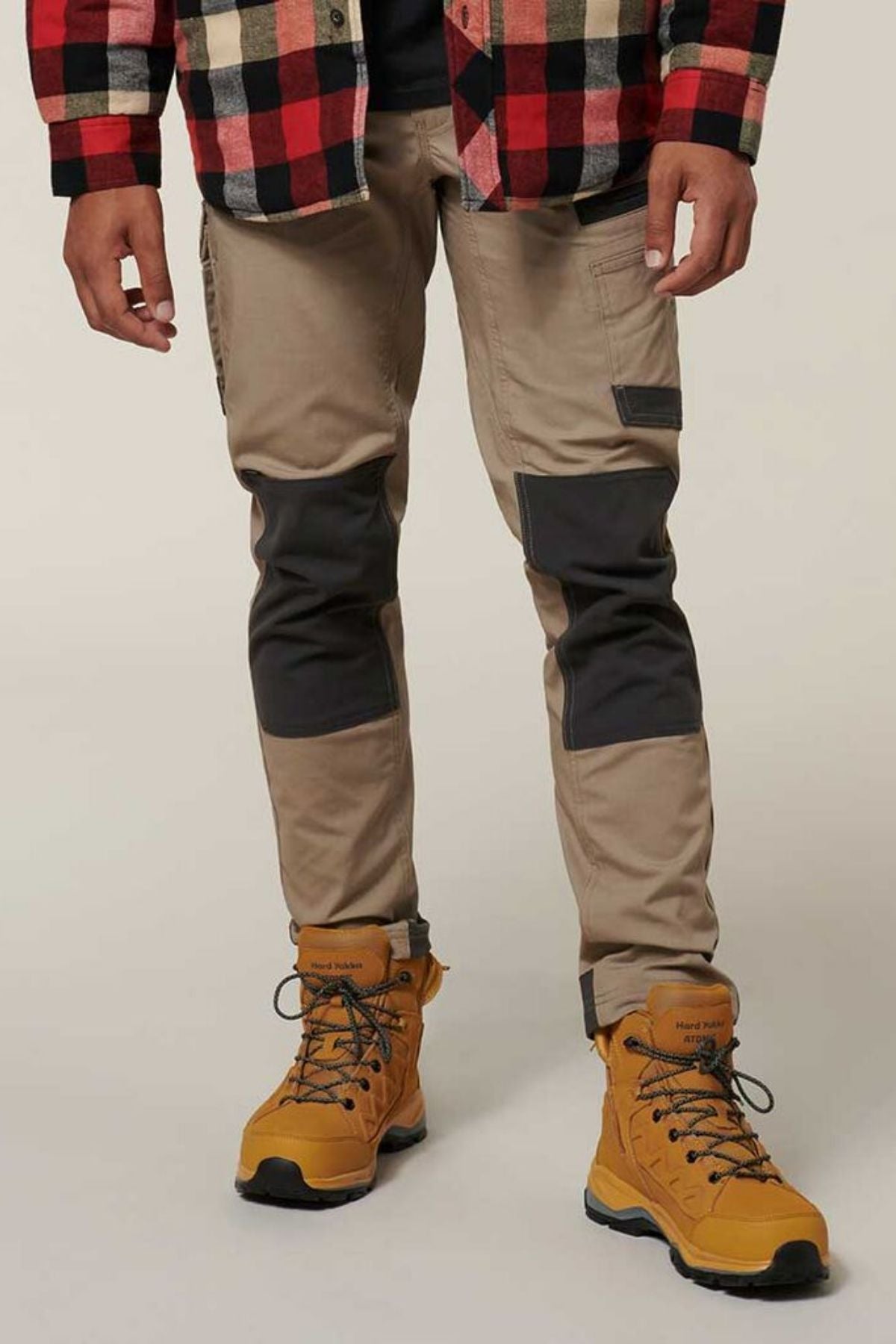 A person is wearing tan Hard Yakka Raptor Pants with reinforced knee patches, a red and black plaid shirt, and yellow work boots. The image is cropped to show only the lower half of the body against a plain background.