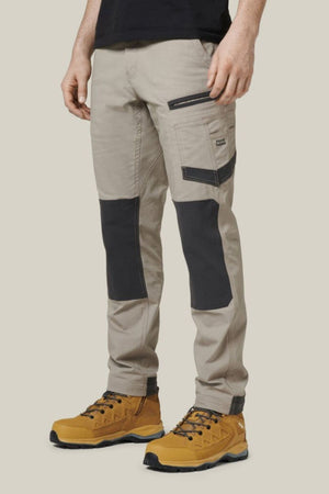 A person wearing Hard Yakka Raptor Pants with Repreve ECO Ripstop Technology, distinguished by their black knee patches and numerous pockets, along with mustard yellow lace-up work boots, stands against a plain light background. The upper body is cropped out of the image.