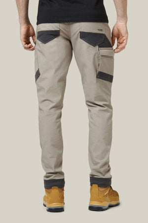 A person wearing beige Hard Yakka Raptor Pants with black pockets and mustard-yellow work boots is standing with their back facing the camera. The pants, made with Repreve ECO Ripstop Technology, feature multiple utility pockets on the sides and back. They are also wearing a black shirt.