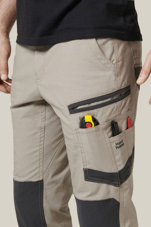 A close-up of a person wearing beige Hard Yakka Raptor Pant with black knee patches and multiple pockets. The pants feature Repreve ECO Ripstop Technology for durability. The pockets contain a utility knife, pen, and other tools, with the brand name "Hard Yakka" visible on one of them.