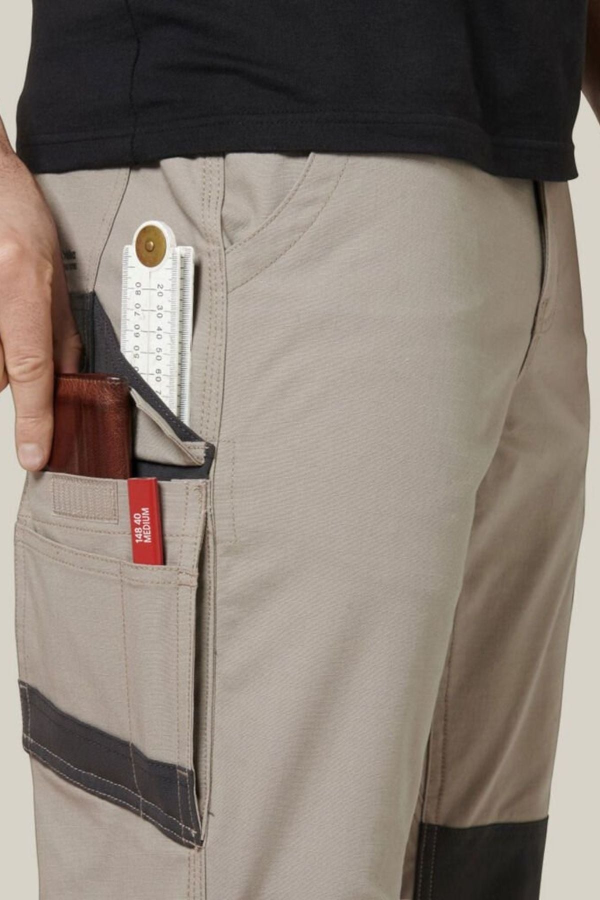 Person wearing Hard Yakka Raptor Pant in beige with black knee patches and a black shirt. They have a folding ruler and a brown wallet partially visible in the side pocket of their pants. The background is plain light grey.