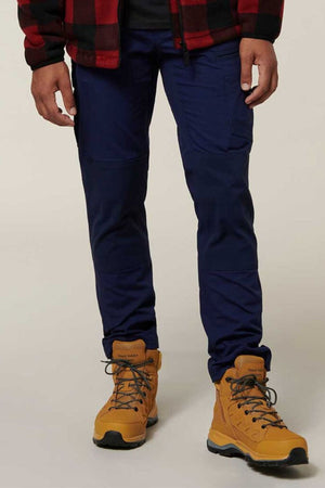 A person is standing against a plain background wearing a red and black plaid jacket, Hard Yakka Raptor Pant in navy blue with reinforced knees, and yellow hiking boots with gray laces. The photo is cropped to show the person from the shoulders down to the feet.