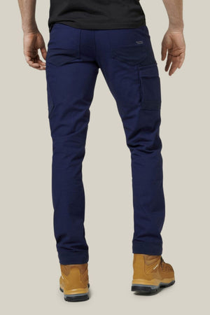 A person wearing Hard Yakka Raptor Pants in navy blue with reinforced knees and tan work boots stands with their back facing the camera. Their hands are relaxed by their sides. The background is plain and neutral.