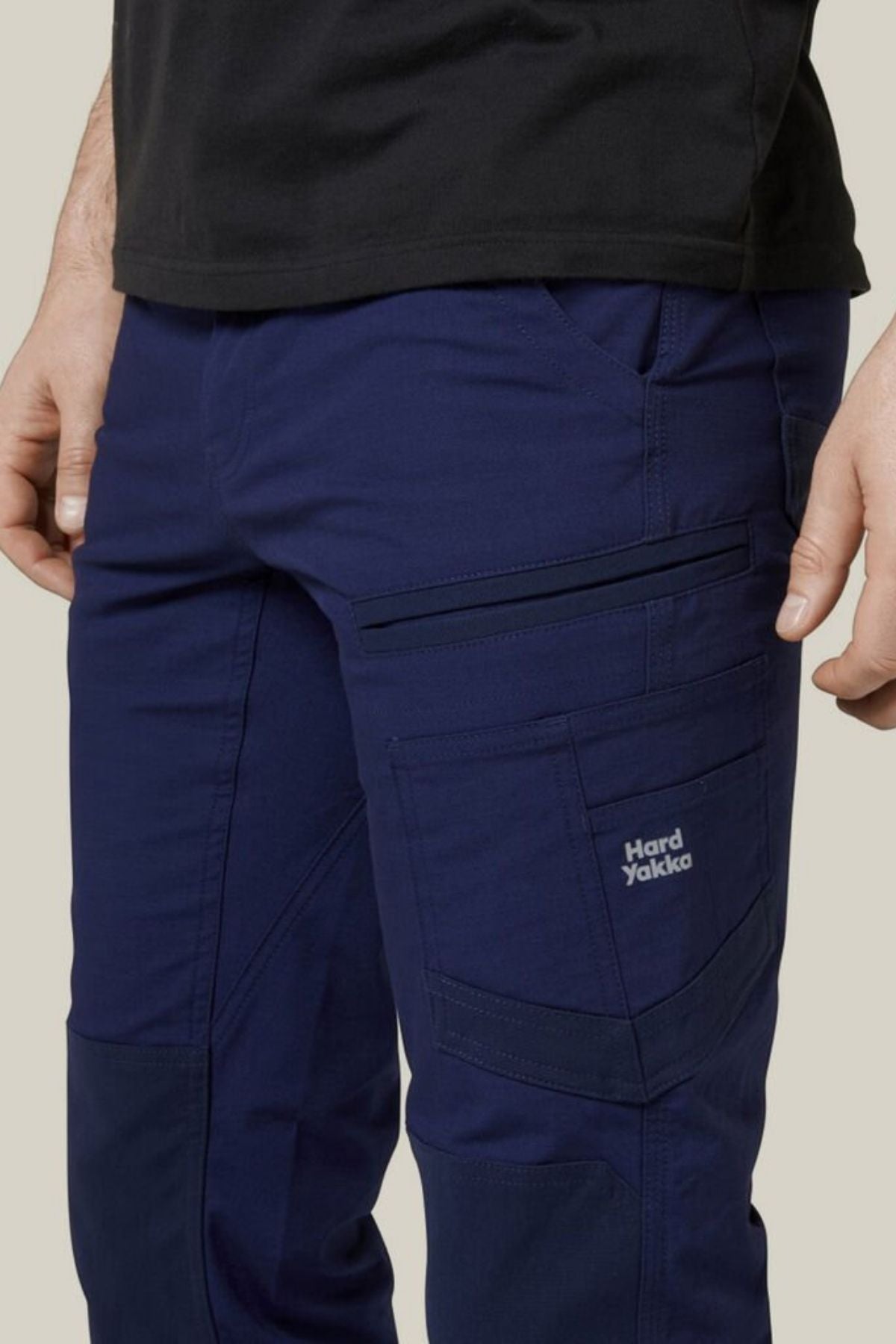 An individual is wearing Hard Yakka Raptor Pants in dark blue, which feature multiple pockets and a "Hard Yakka" logo on the thigh. These pants are designed with reinforced knees and ECO Ripstop Technology for enhanced durability. The person is also donning a black shirt, set against a plain beige background.