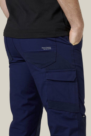 A person wearing Hard Yakka Raptor Pants in navy blue, featuring reinforced knees and a visible "Hard Yakka" logo on the back pocket. The work pant includes multiple large pockets, with one of them holding their hand. They are also wearing a black top, showcasing both style and functionality.