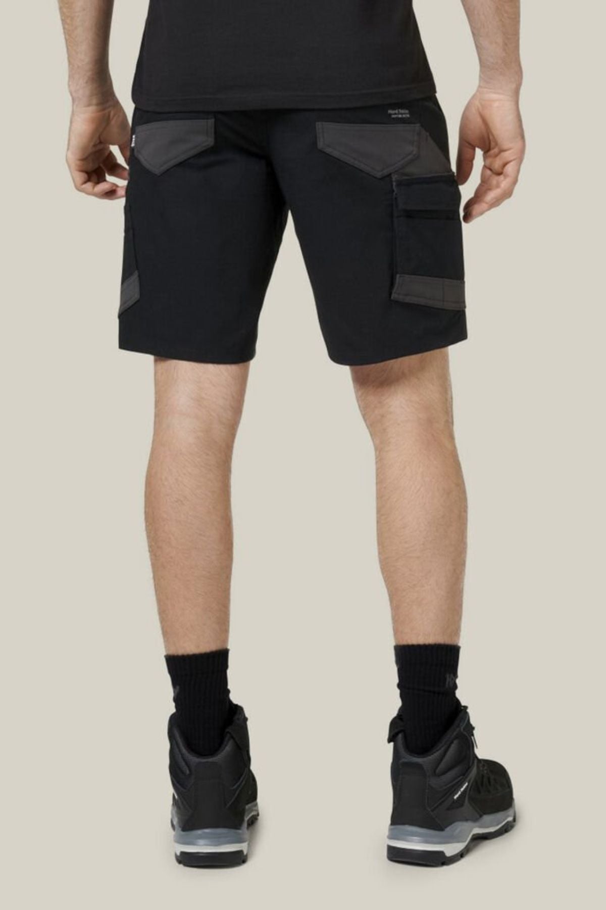A person wears Hard Yakka Raptor Mid Shorts with an Active Fit stretch waistband and dark gray pockets, a black shirt, and black sneakers with white soles. They stand on a light gray background, facing away from the camera.