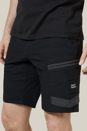 A person is wearing black Hard Yakka Raptor Mid Shorts with side pockets and iconic logo, paired with a black shirt. These casual-fit shorts feature an Active Fit stretch waistband and comfort-enhancing Repreve ECO Ripstop Technology, focusing on the lower torso and upper legs.