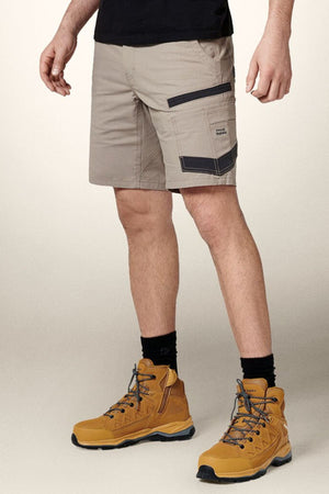 A person is wearing Hard Yakka Raptor Mid Shorts in beige, crafted from durable stretch ripstop fabric with black accents and pockets, paired with black socks and tan hiking boots. The upper part of the body is not visible, set against a plain, light-colored wall.
