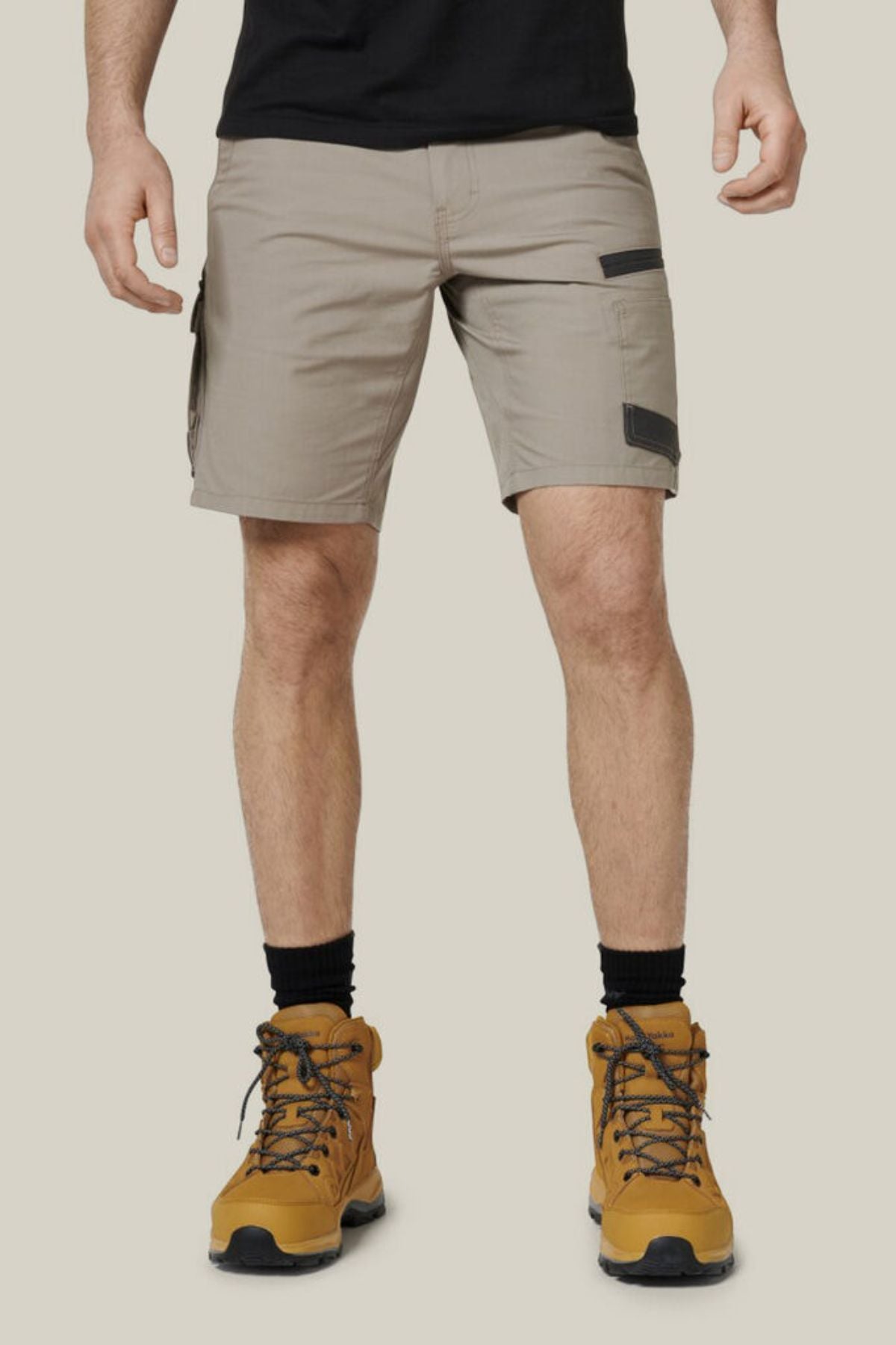 A person wearing the Hard Yakka Raptor Mid Shorts in light gray, made from durable stretch ripstop fabric, and black socks stands against a plain, light beige background. They are also wearing yellow-brown lace-up hiking boots. The person's head is not visible in the image.