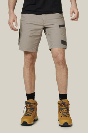 A person wearing the Hard Yakka Raptor Mid Shorts in light gray, made from durable stretch ripstop fabric, and black socks stands against a plain, light beige background. They are also wearing yellow-brown lace-up hiking boots. The person's head is not visible in the image.
