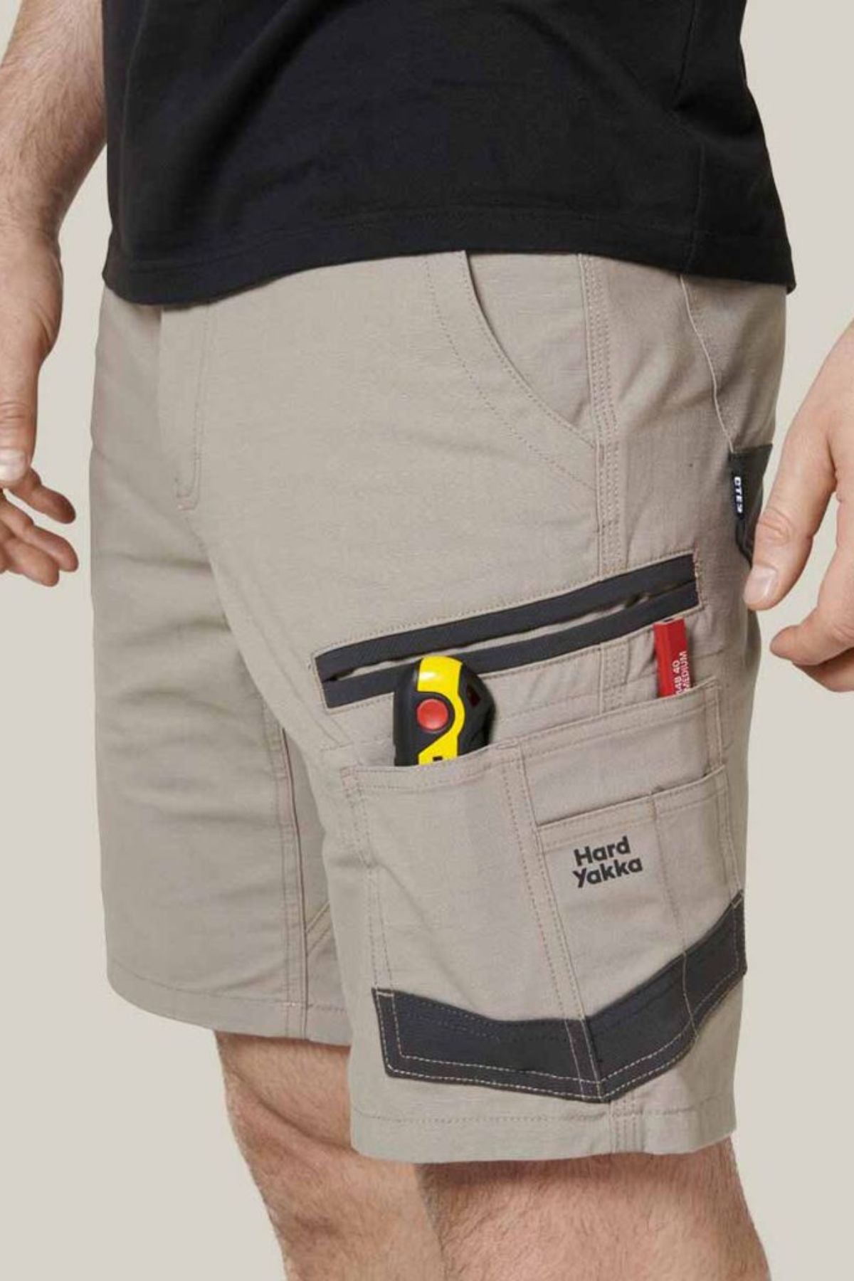 A person wearing the Hard Yakka Raptor Mid Shorts, featuring beige color with black accents and multiple pockets, including one holding a yellow utility knife. The words "Hard Yakka" are emblazoned on one of the pockets. The shorts also feature an Active Fit stretch waistband and are paired with a black shirt against a plain beige background.