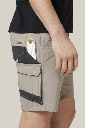 A person wearing Hard Yakka Raptor Mid Shorts in beige with multiple pockets, including a cargo pocket securing a measuring tape. These utility shorts feature an Active Fit stretch waistband for comfort. The person is wearing a black shirt, with only their lower torso and right hand visible against a plain, light-colored background.