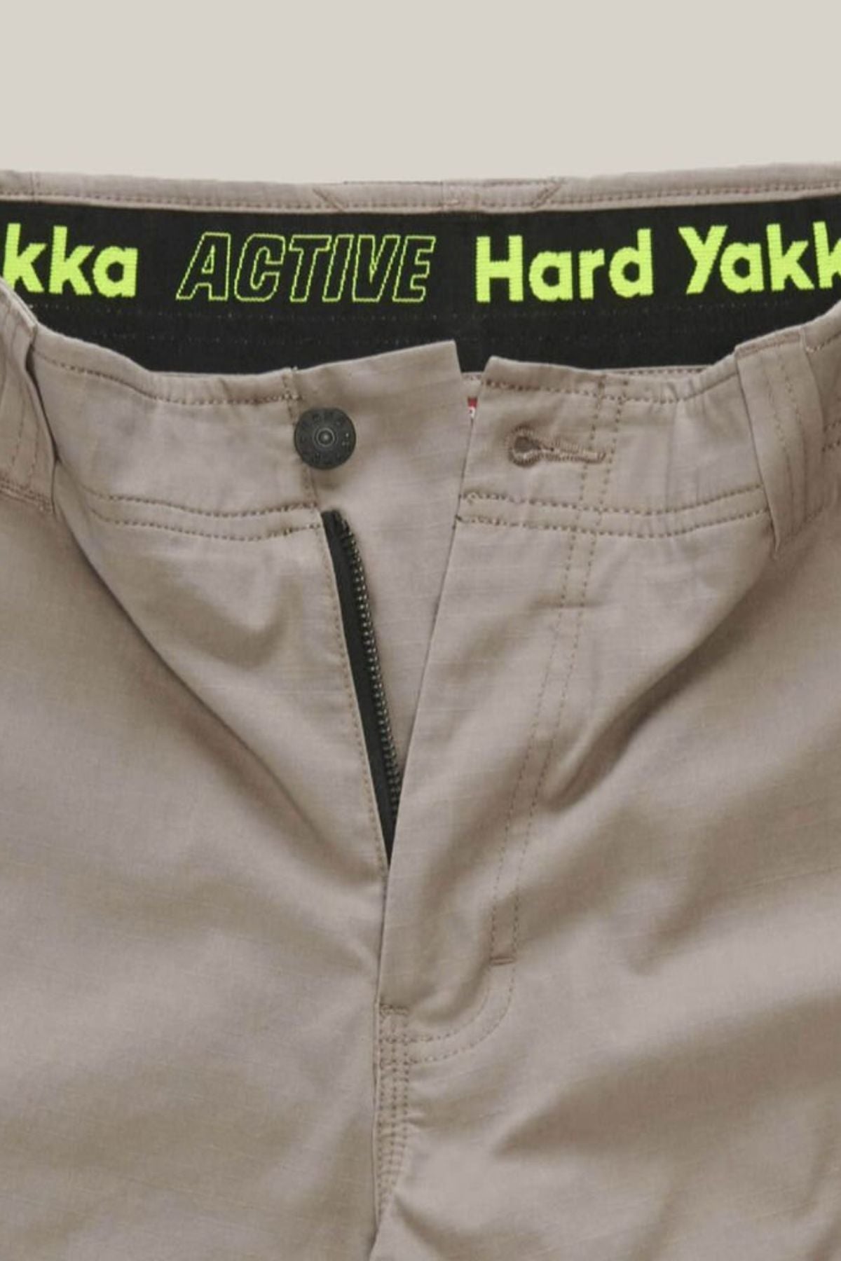 Beige Hard Yakka Raptor Mid Shorts with the zipper partially open, featuring a black waistband with green text that reads "Hard Yakka ACTIVE," made from durable stretch ripstop fabric.