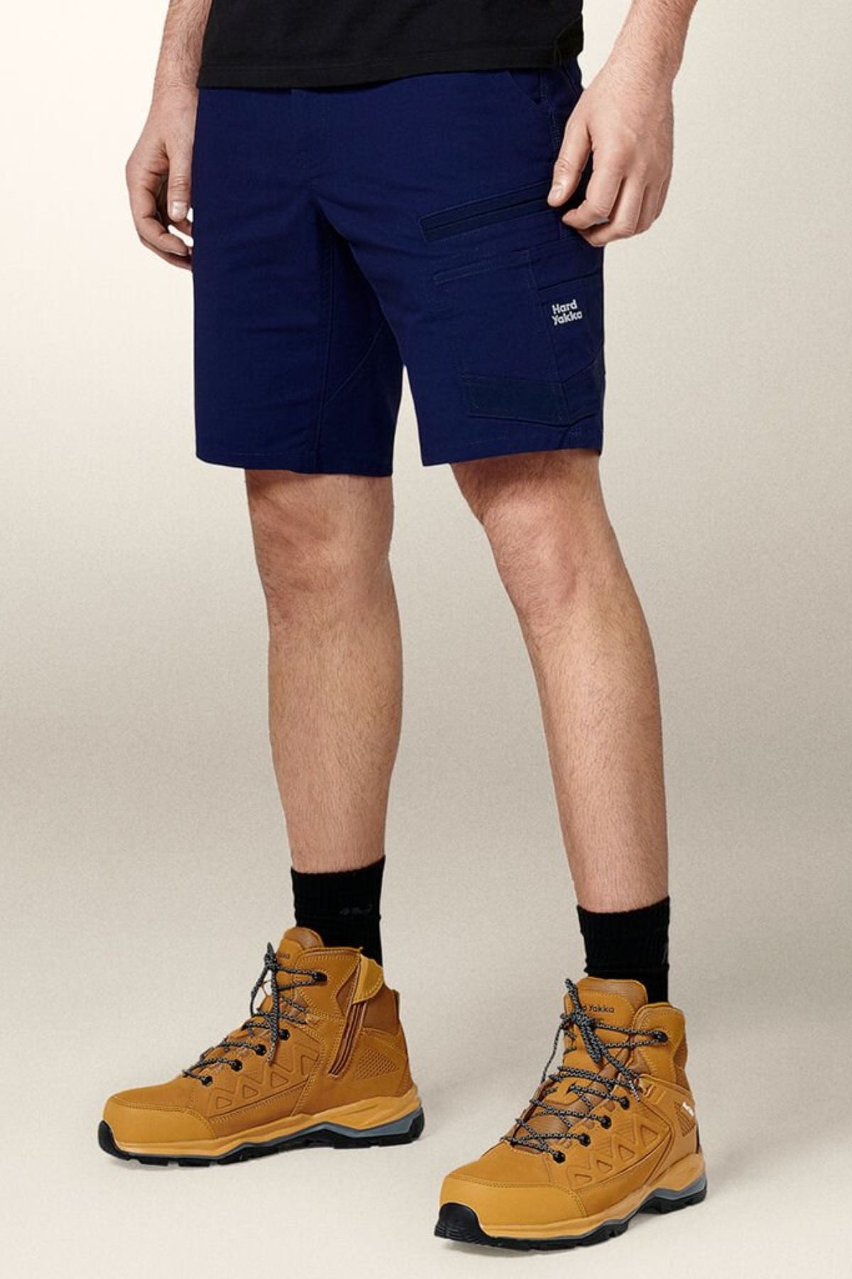 A person wears Hard Yakka Raptor Mid Shorts in navy with an Active Fit stretch waistband, zipper pockets, small white logo on the left thigh, and Repreve ECO Ripstop Technology, standing against a neutral background.
