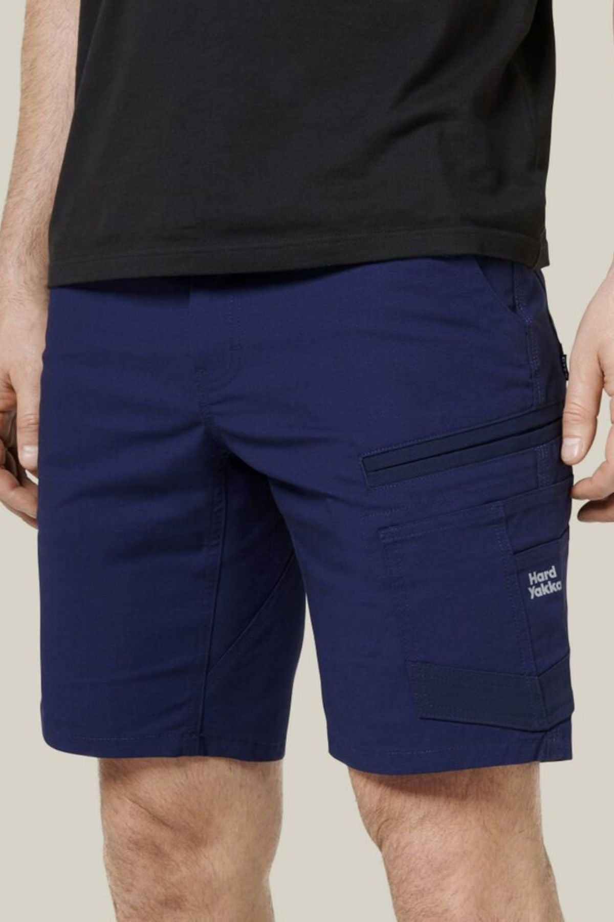 A person wears a black t-shirt and Hard Yakka Raptor Mid Short in navy blue, with multiple pockets (including a zippered one), an Active Fit stretch waistband, and a small "Hard Yakka" logo on the left leg. Shorts shown waist to mid-thigh.