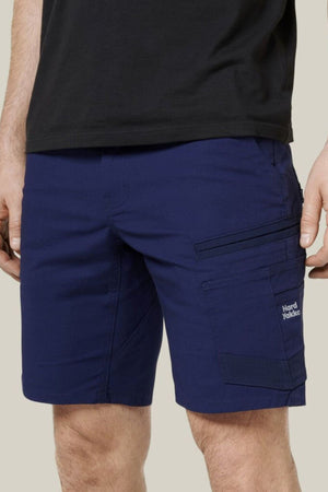 A person wears a black t-shirt and Hard Yakka Raptor Mid Short in navy blue, with multiple pockets (including a zippered one), an Active Fit stretch waistband, and a small "Hard Yakka" logo on the left leg. Shorts shown waist to mid-thigh.