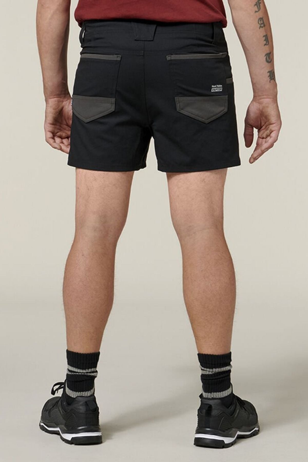 A person is shown from the back wearing Hard Yakka Raptor Short Shorts in black, featuring Sustainable REPREVEĀ® ECO Ripstop Technology with dark gray pocket accents. They are also donning a red shirt, black socks, and black sneakers. The person's right arm is adorned with tattoos as they stand against a plain, light-colored background.